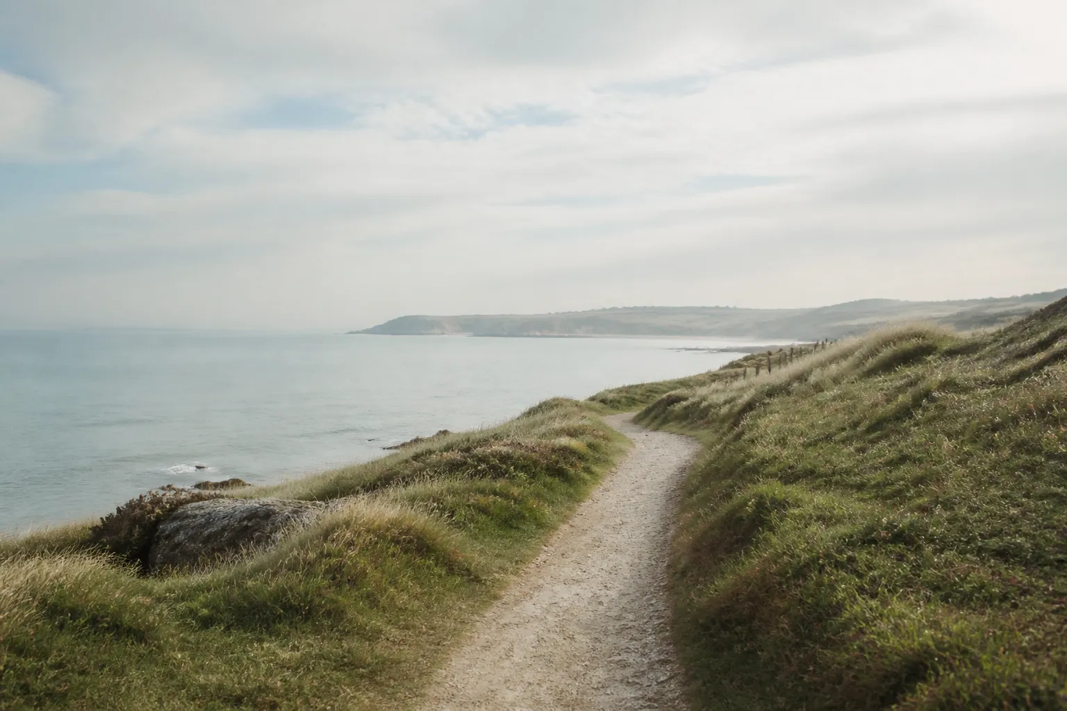 A quiet coastal path curving through grassy dunes towards a calm, pale sea
